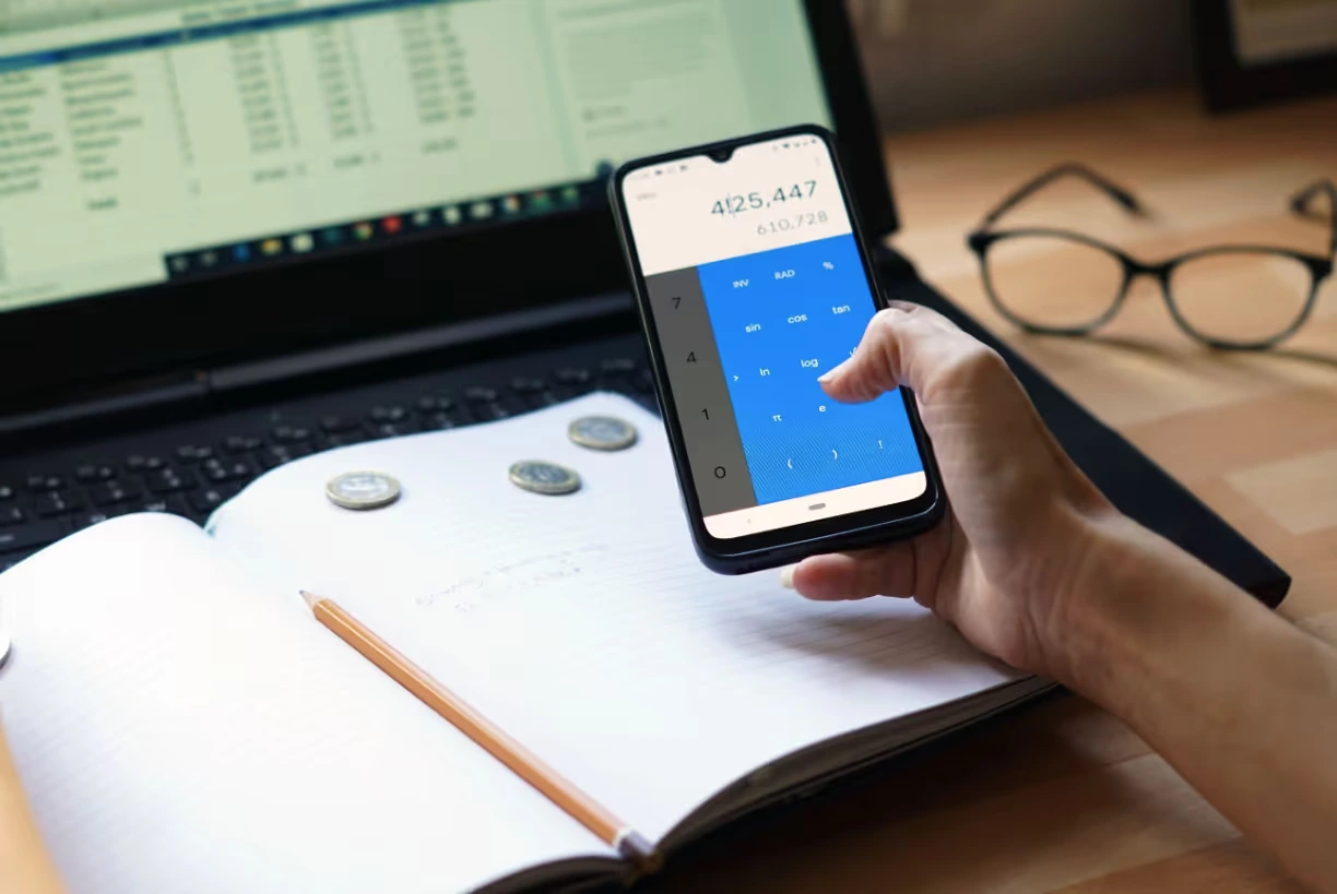A hand working a calculator app on a phone over an open notebook, beside a laptop showing a spreadsheet